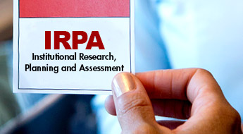 !A woman’s hand holding a card with red letters reading “IRPA Institutional Research, Planning and Assessment.”