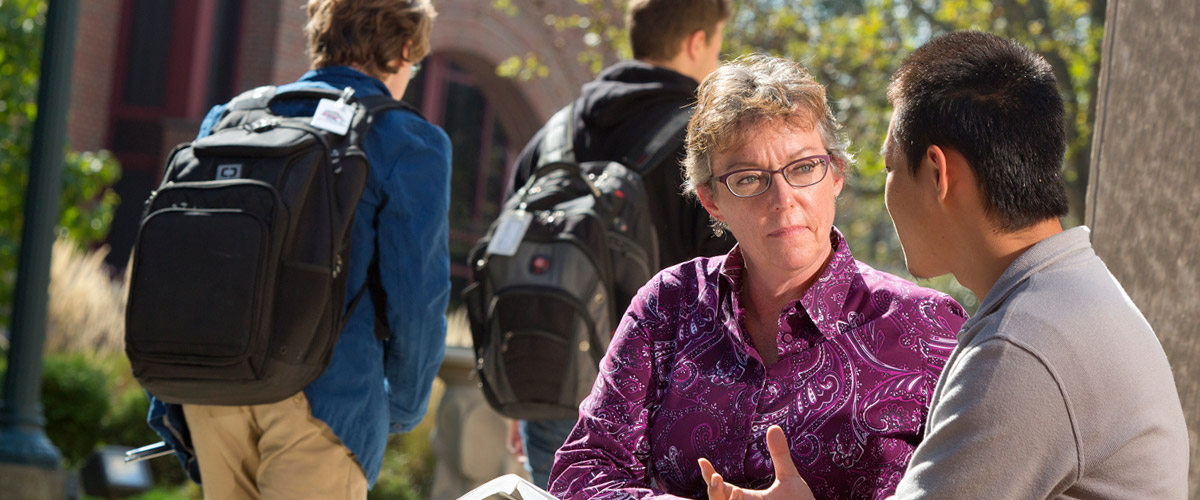 Professor Julia Williams sits outdoors on campus taking with another member of the campus community as students walk in the background to their classes