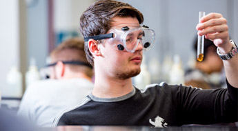 Male student wearing goggles examines test tube. 