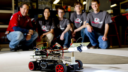 Small, wheeled robot in the foreground with robotics team and instructor kneel in the background.