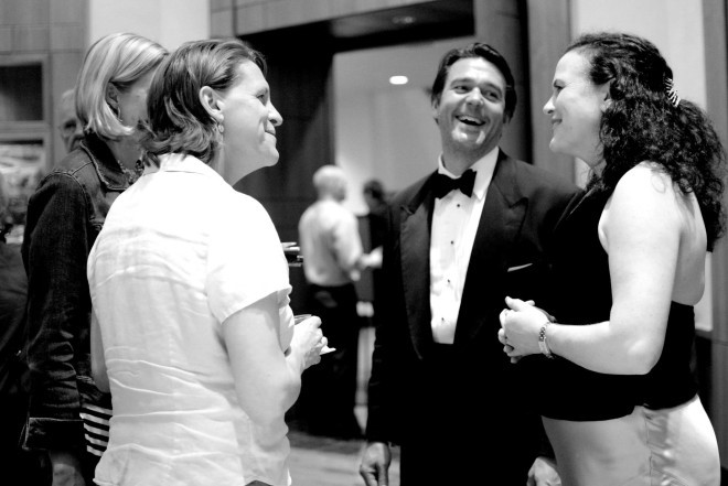 Black and white photo shows patrons of Hatfield Hall’s theater in the lobby of Hatfield Hall talking and smiling with each other. 