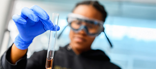 A student wearing gloves and safety goggles holds up a test tube to examine its contents.