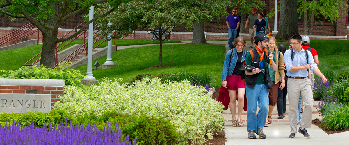 Image shows students walking through Root Quadrangle amid green bushes, purple flowers, and green grass.