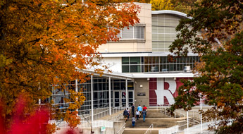 !Image shows students walking up the front steps of the Sports and Recreation Center where a big Rose-Hulman R is on the exterior wall. The image is framed on the right and left by colorful leaves of two trees.