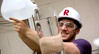 Male student wearing hardhat pours water into a vessel.