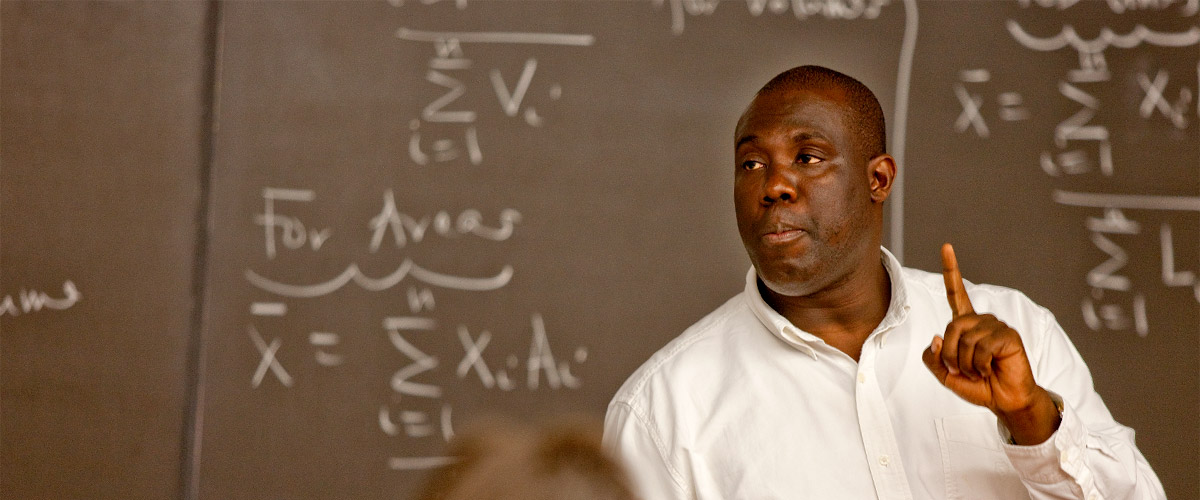 Image shows a professor lecturing in front of a chalk board covered in sophisticated math equations. 