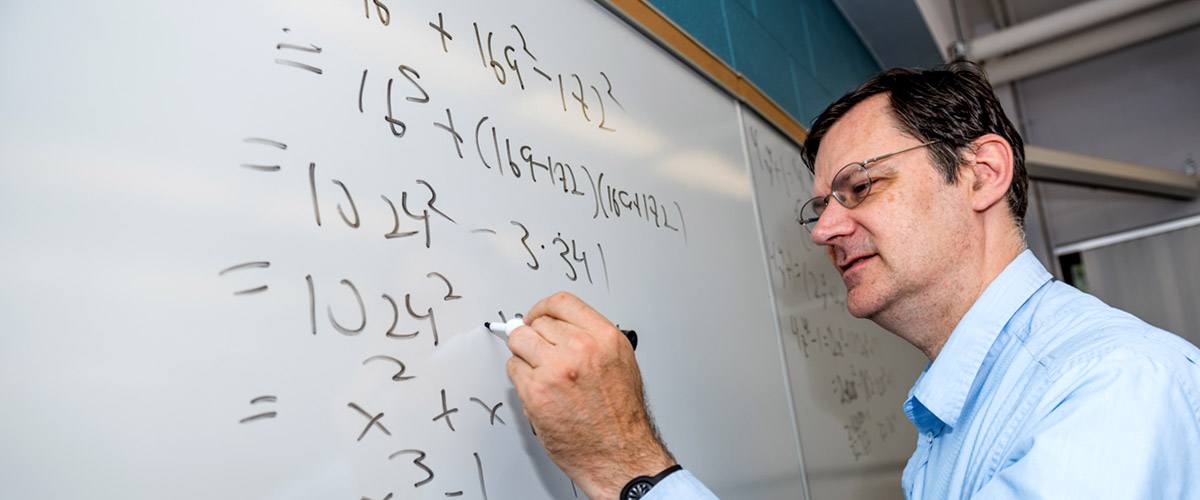 Professor John Rickert writes math equations on a white board while teaching a class.