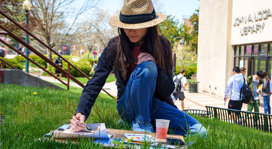 !Female student sit in the grass in Root Quadrangle doing watercolor drawing.