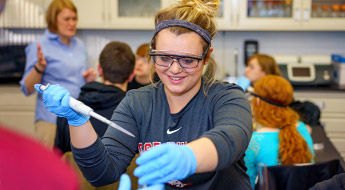 Several students and a professor watch as a student performs an experiment in a campus laboratory.