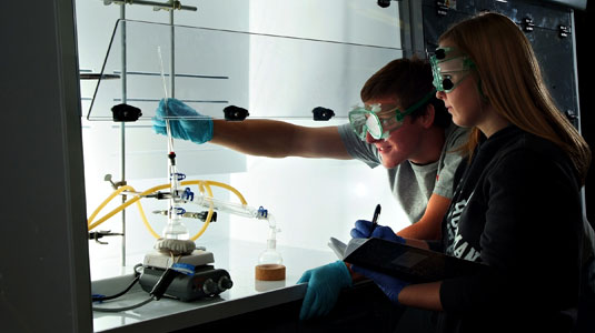 Male and female students wearing goggles perform an experiment in chemistry lab.