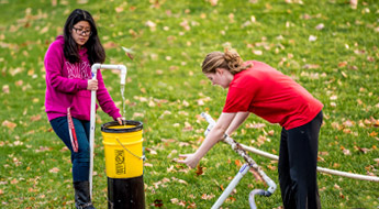 Two female students work with PVC pipes and buckets outside.