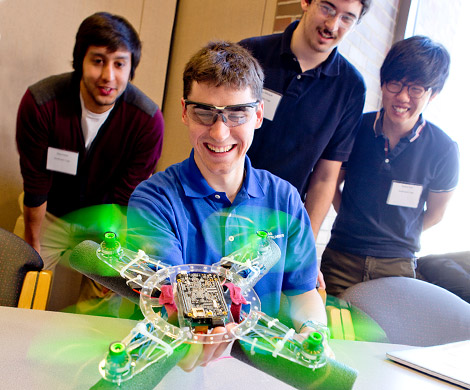Male student holds drone with green propellers spinning as three other male students look on. 