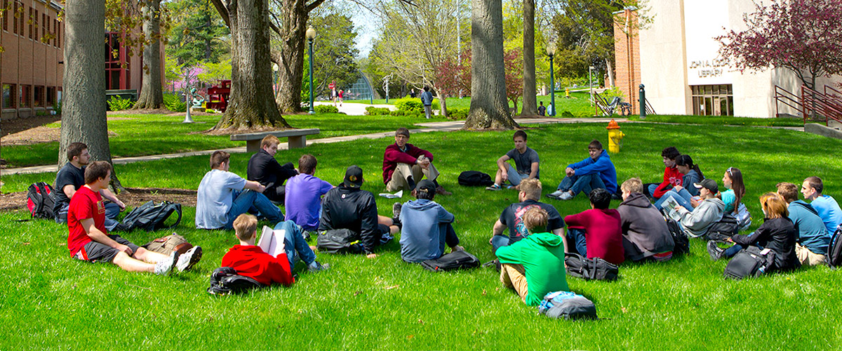 A group of about 20 students seated in the grass on campus listening to a lecture. 