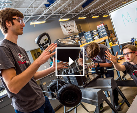 A male student tosses a small wheel and tire to another male student in a workshop setting.