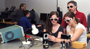 Two female students work with optics devices in a laboratory as male professor looks on.