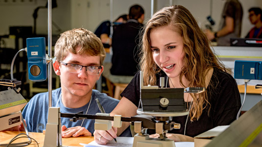 Male student & female student use a scale in a physics lab.