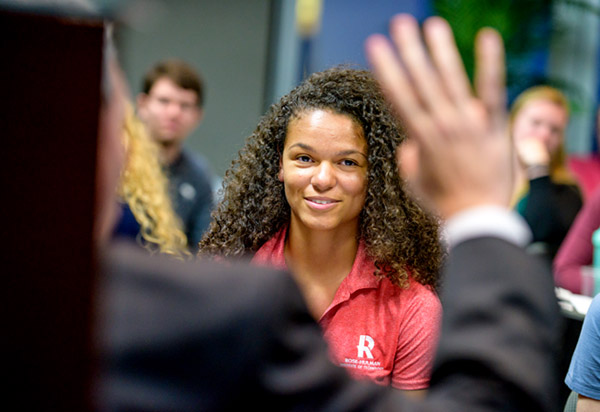 African American female among others in  a workshop with their hands raised as if to ask a question