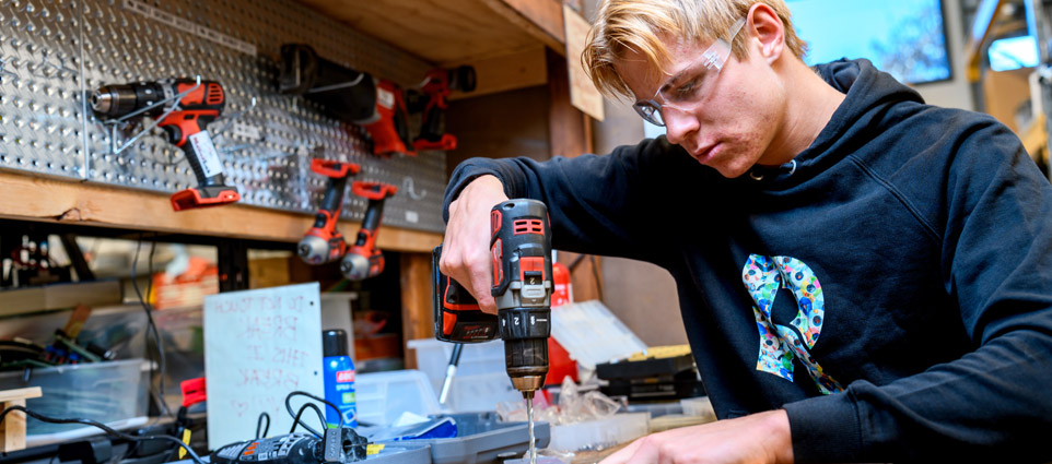 A student wearing safety goggles uses a drill and woodworking equipment.