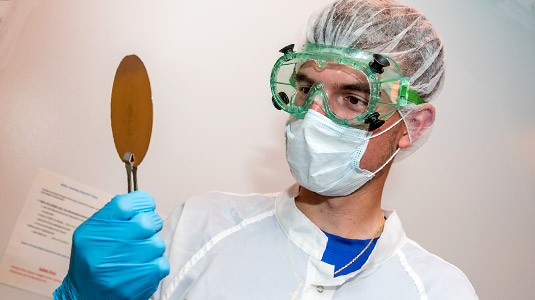 Male in goggles, mask, and gloves examines a metal disc.