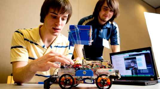 Two male students make adjustments to a small wheeled robot.