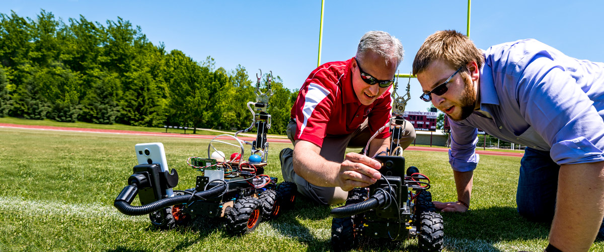 Dr. David Fisher and a male student kneel on a grassy field to make adjustments to a robotic vehicle.