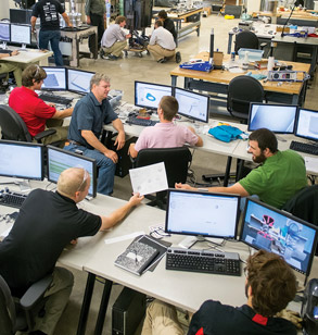 !Group of students talk while sitting at their desks with computers