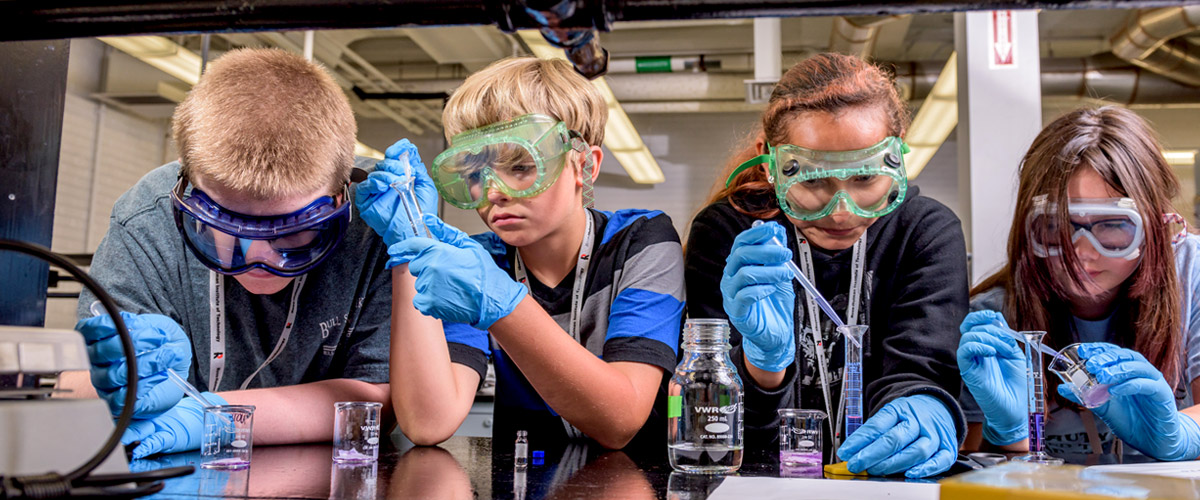 Four young students wearing lab gloves and goggles work on a laboratory table