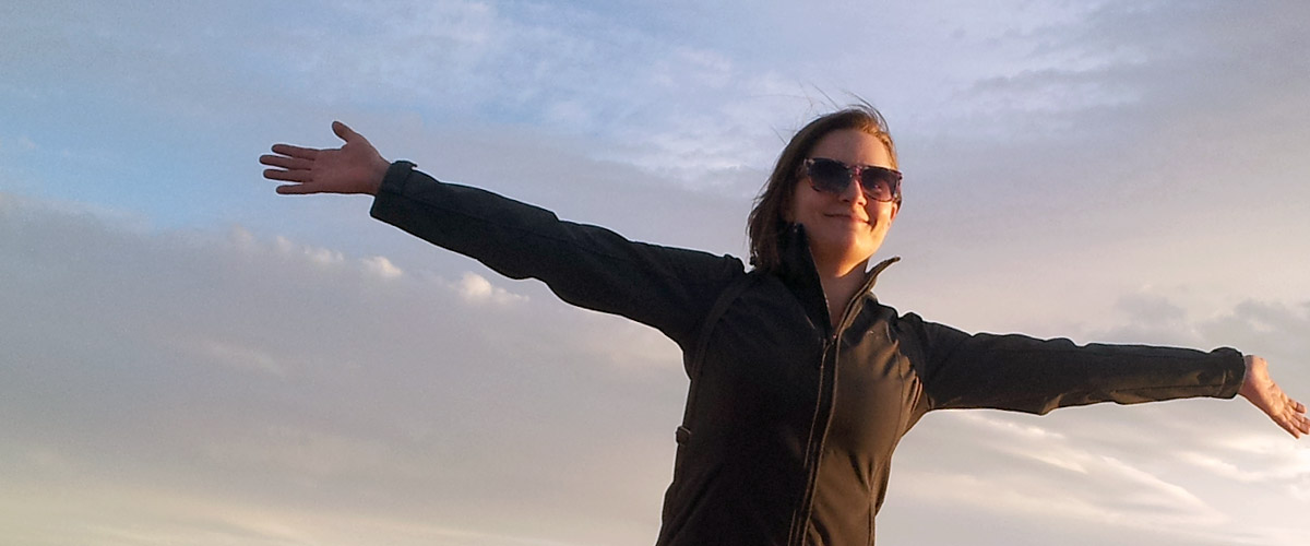 Smiling female student holding her arms out to her sides against a beautiful sky at dusk.