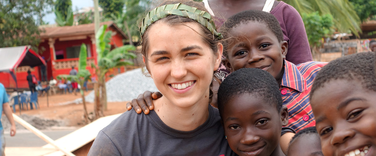 Female student wearing paper crown of leaves, hugging children in a village in Ghana.