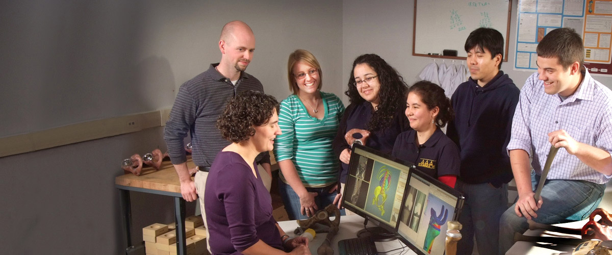 A small group of students smiling and looking at Professor Renee Rogge as she sits at her desk discussing computer images of joint replacement devices