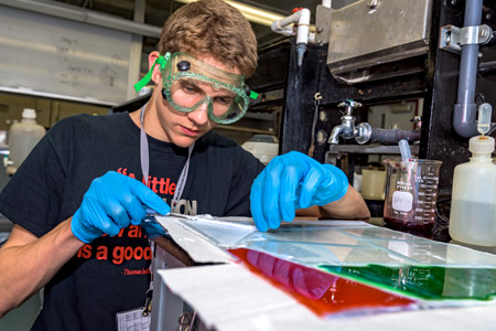 A male Catapult student wearing goggles and blue latex gloves while using a machine to carefully cut pieces of clear plastic.