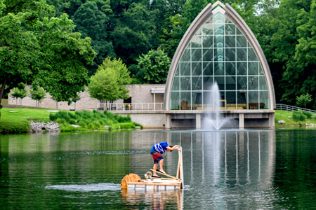 A male Catapult student operating a walk-on-water device in the middle of Speed Lake with White Chapel and the lake’s fountain in the background.
