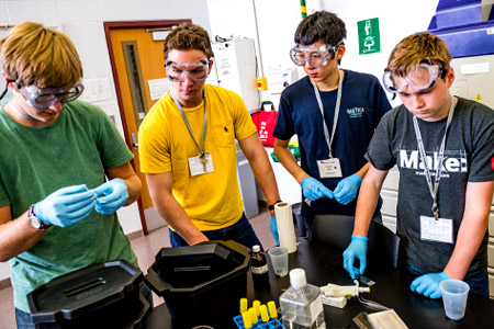 A group of male Catapult students wearing goggles and latex gloves working together on a project.