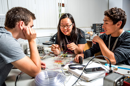 Two male and one female Catapult students smiling while working together on a project involving electrical wiring and plastic tubing. 