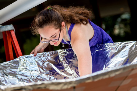 Female Catapult student working on a large, silver solar oven.