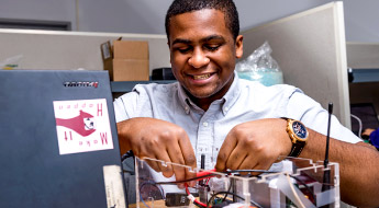 !Image shows a male student working on an electronics project and smiling in a lab on campus.
