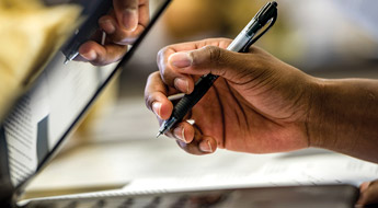 !Image shows a student’s hand holding a pen next to a laptop computer. The student is taking notes while working.