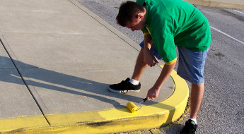 !Image shows a male student using a paint roller to paint a yellow “no parking” curb on a street somewhere on campus.