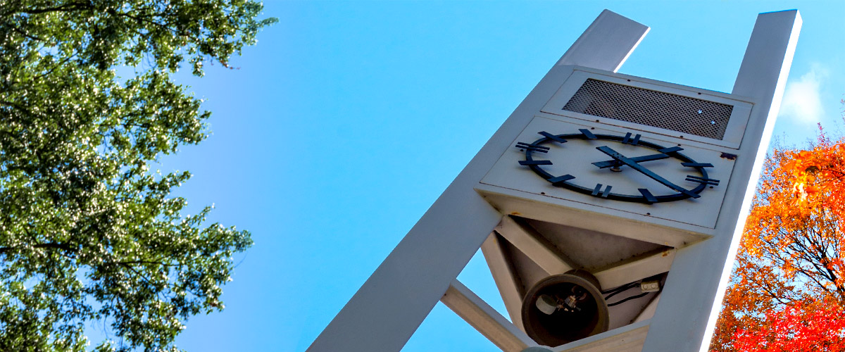 Image shows the carillon clock tower that is outside of Hadley Hall at the main entrance to Rose-Hulman. It is flanked by a green tree on one side and a tree with orange fall leaves on the other. It stands in front of a clear blue sky