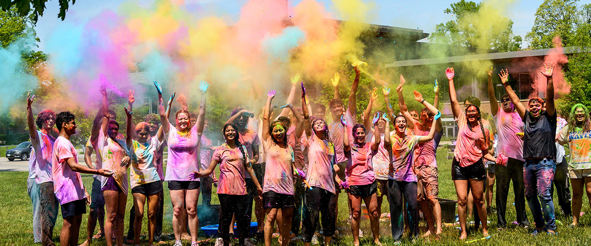 A group of students stand outside covered in colored powder while they celebrate Holi festival of colors.
