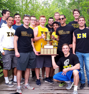 Members of Sigma Nu pose with a large trophy outside in a wooded area of campus. 