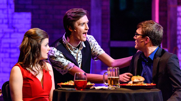 A male actor gestures to another male actor seated at a dining table as female actor looks on.