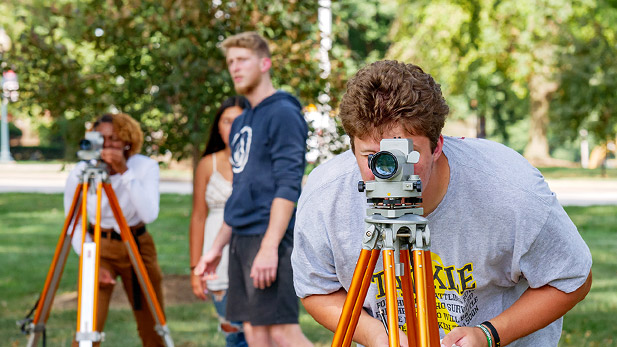 Civil engineering students peer through the lens of surveying equipment on tripods outside on campus.