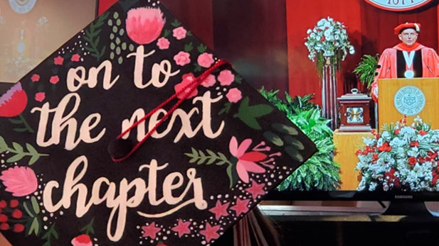 Image shows top of a decorated student graduation cap reading: "The Next Chapter Begins." President Coons in regalia is at the podium in distance.