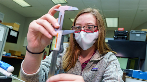 Female student intern using measuring device working on a project. She is wearing a mask to prevent spread of COVID-19.