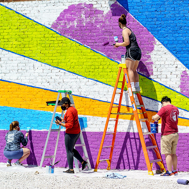 Students painting a colorful mural on the side of a large brick building.