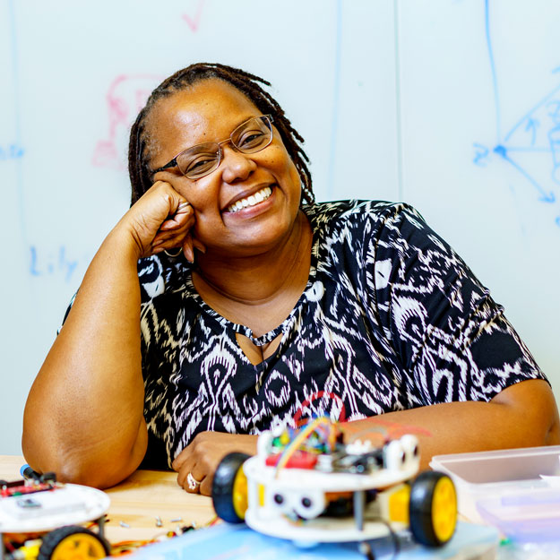 Carlotta Berry smiles behind a table covered with robots and wires. 