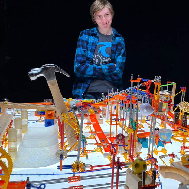 Gabe Neise stands proudly behind one of his machines, which contains several hammers and billiards balls, K'NEX pieces, dominoes, and other small household objects.