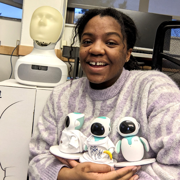 Left: Nyomi Morris grins and gives two thumbs up next to her research poster. Middle: Nyomi Morris stands next to a banner advertising an event. Top right: Nyomi Morris smiles and holds three small Nao robots to her chest. Bottom right: Nyomi Morris poses with the Duolingo owl mascot. 