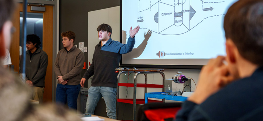 A student points to a PowerPoint slide with the design for the jet engine heat exchanger.